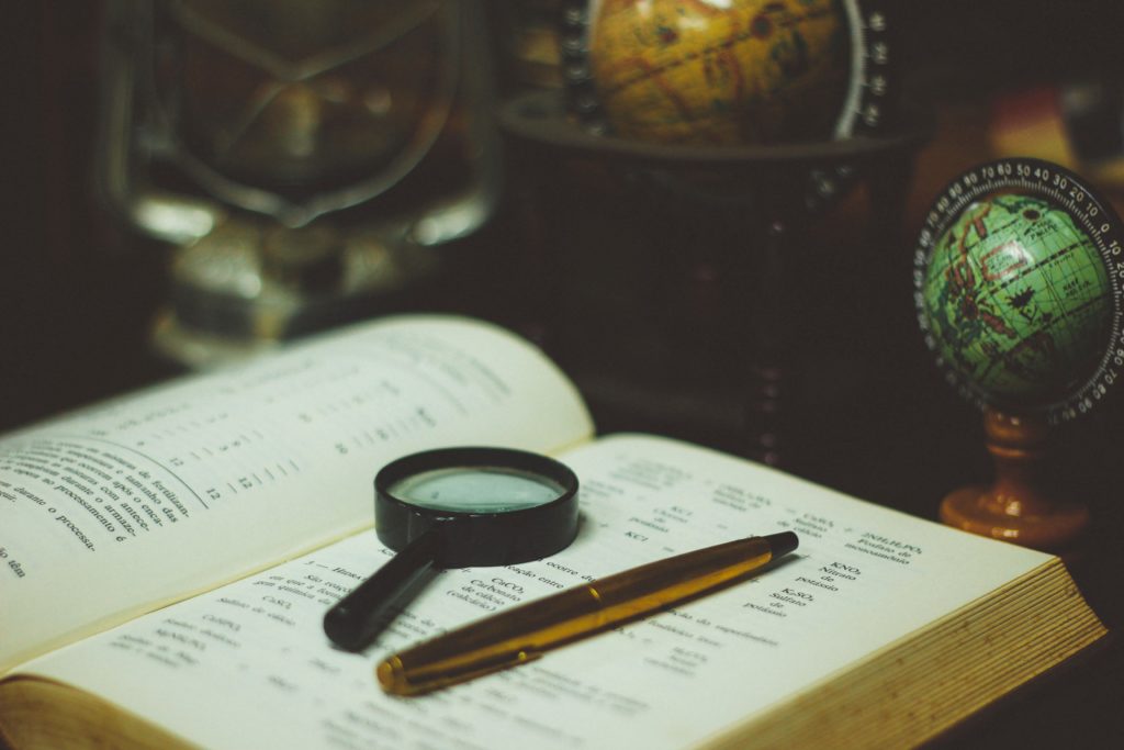 An open book with magnifying glass and pen sitting on top, surrounded by globes