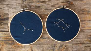 Two embroidery hoops rest side by side on a wooden table. Both have dark blue fabric with embroidered constellations on it.
