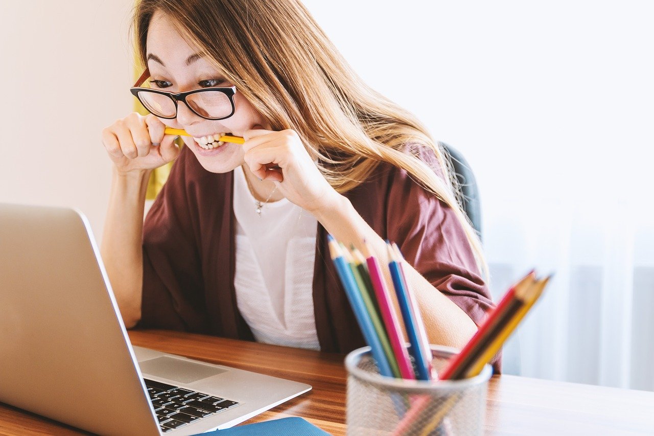 A young lady wearing glasses looking at a laptop while biting a pencil between her teeth in what looks like anxiety.