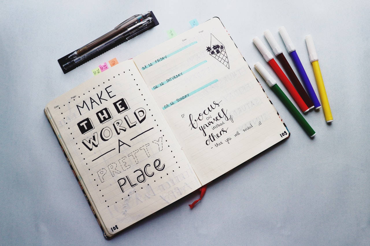 A journal is open on a table. It has artistic writing in it and doodles. There are two pens about the journal and five colorful markers to the right of the journal.