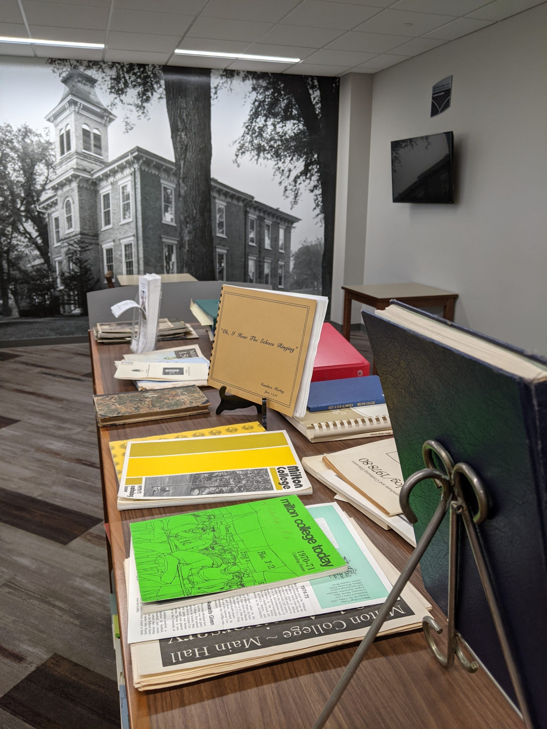 Image of various Milton College paraphenalia displayed atop a book shelf with a mural image of an old campus building.