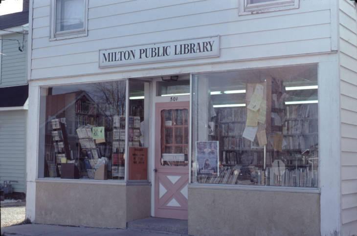 Picture of old library location on West side of town. White building front with large windows and a sign that reads Milton Public Library