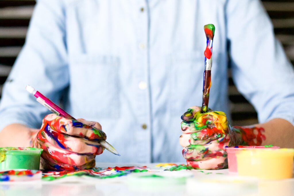A person holding a pencil and a paintbrush with hands full of paint.