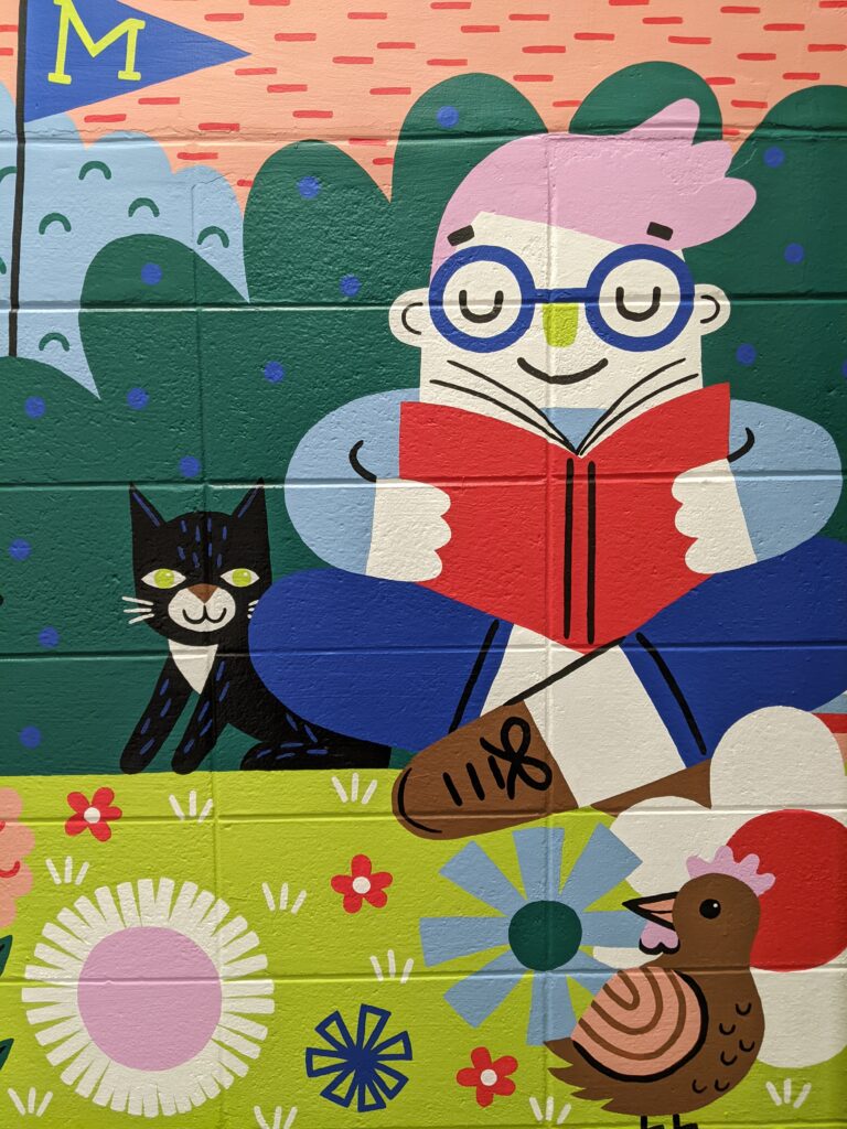 Picture of the mural painted in our stairwell with a child reading a book with a cat, chicken, and flowers around them