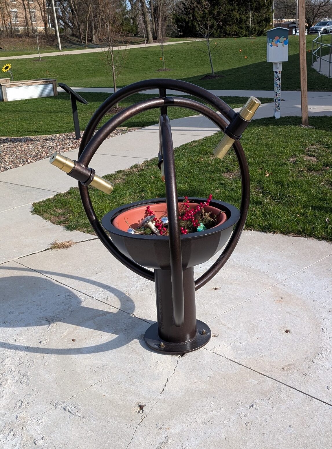 A circular kaleidoscope with metal framework on a concrete path, surrounded by grass and trees in a park setting.