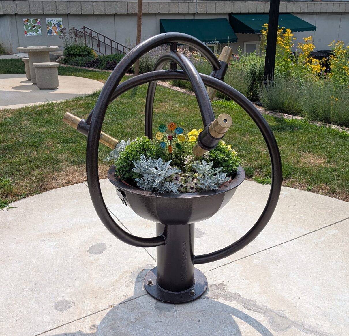 A circular kaleidoscope with metal framework on a concrete path, surrounded by grass and trees in a park setting.