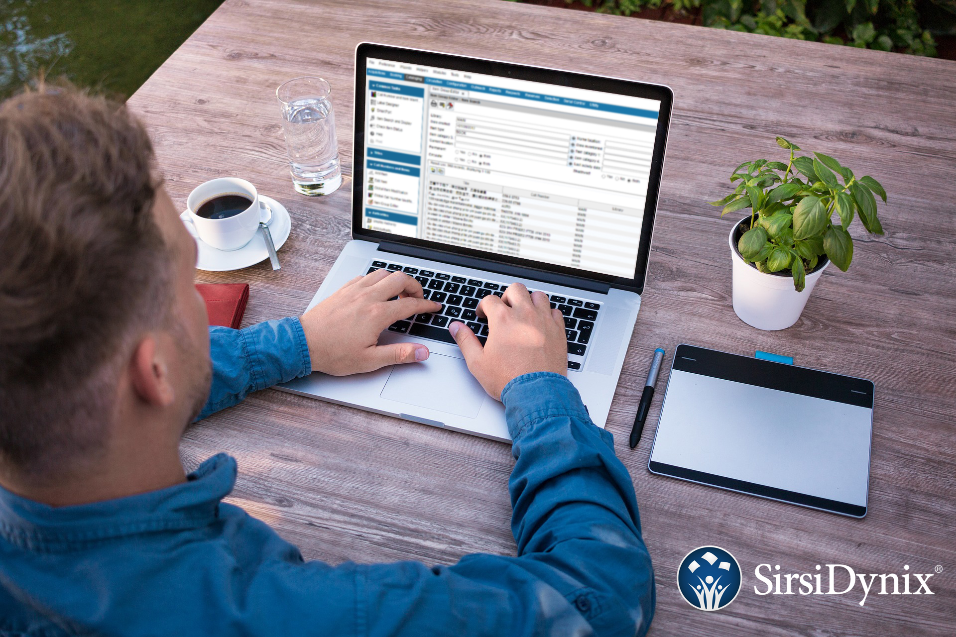 Man in blue shirt sits at a wooden desk. He is working on a laptop displaying a SirsiDynix Workflows window.